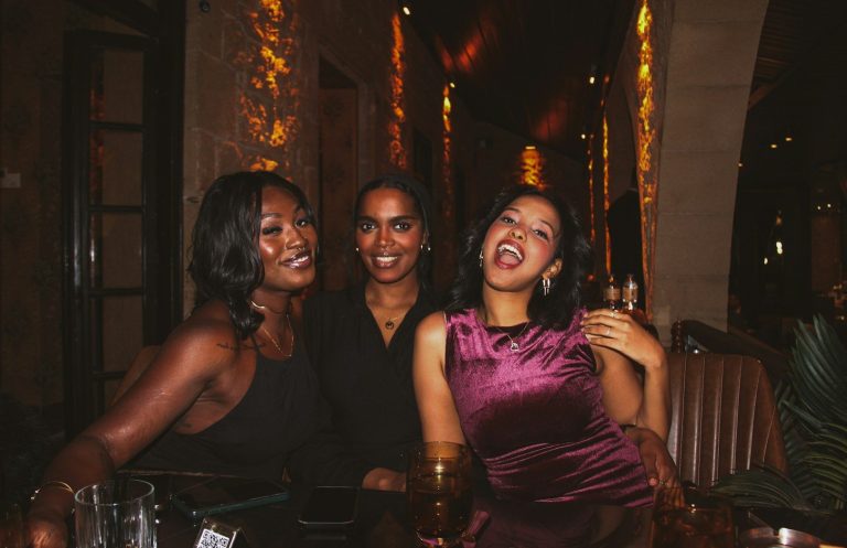 Three friends smiling and posing together in a warmly lit restaurant setting.