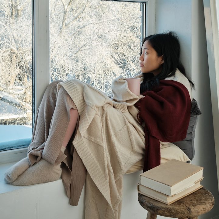 A woman wrapped in blankets sits by a window, gazing thoughtfully outside.