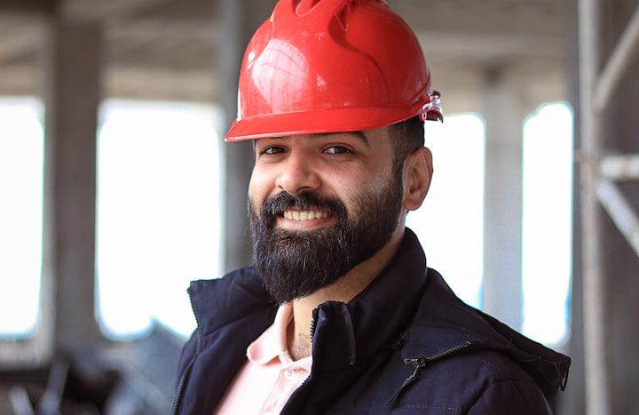 Smiling man in a hard hat on a construction site, wearing a dark jacket over a light shirt.