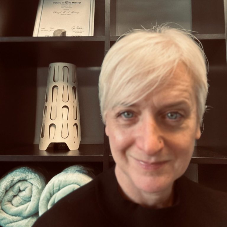 A smiling massage therapist with short blond hair, sitting in a massage treatment room.