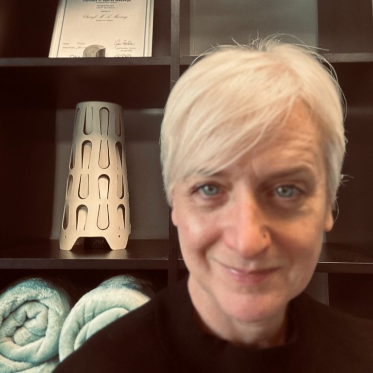 A massage therapist with short blond hair smiles in her treatment room.