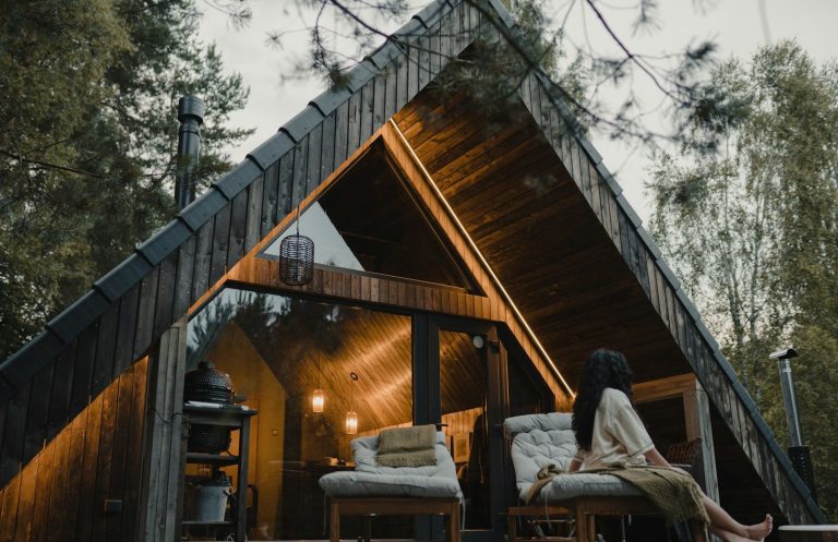 A wooden A-frame holiday cabin with warm lighting, featuring seating and a person relaxing outside.